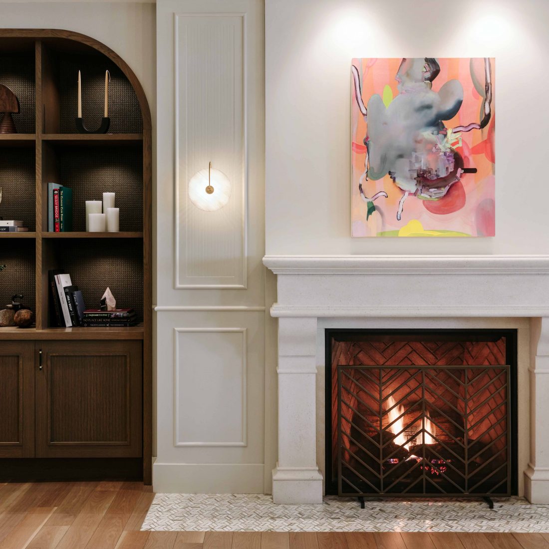 Brown bookshelf filled with trinkets to the left of a burning fire place with a colourful painting above fireplace