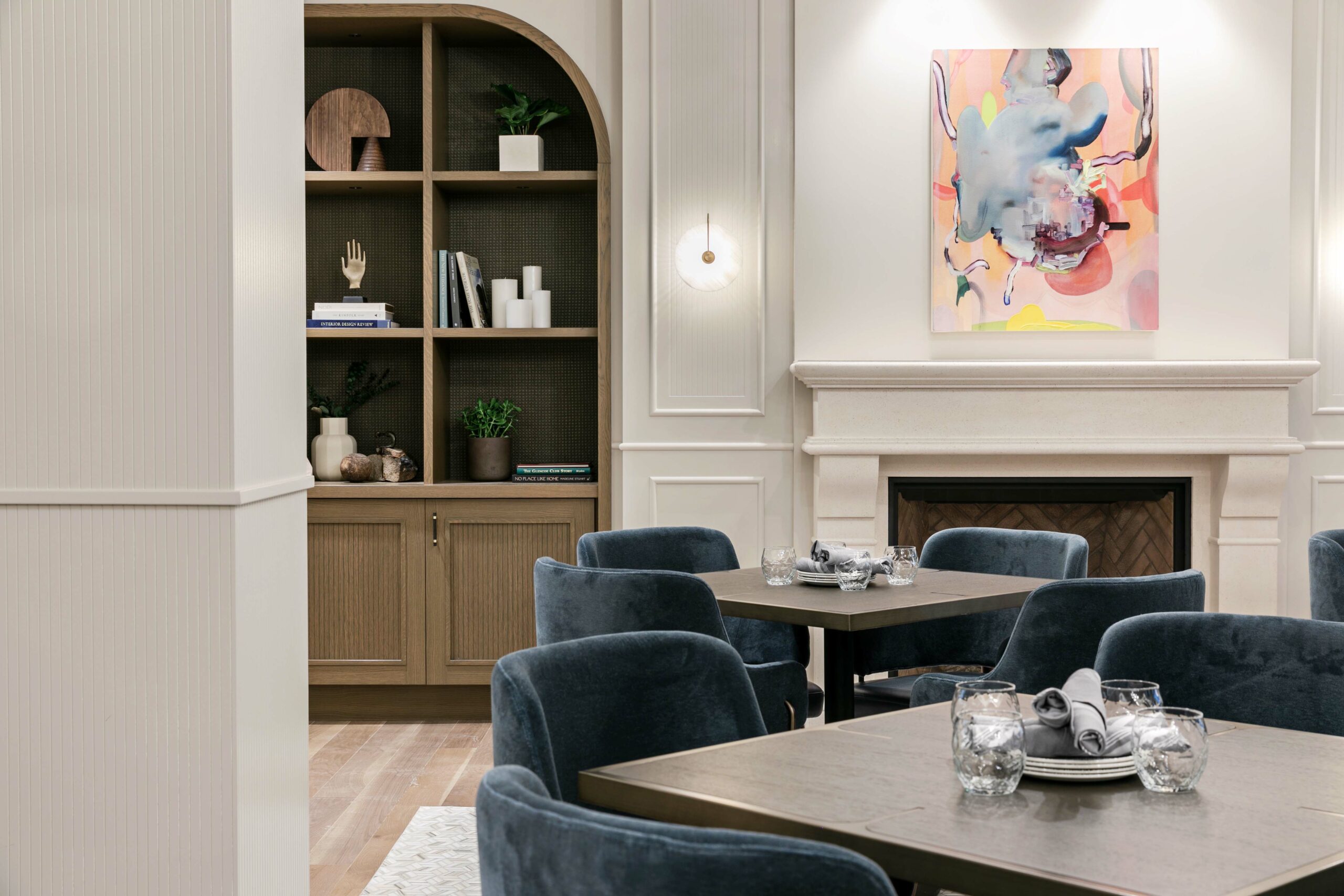 Big blue fabric chairs facing set dining tables, colourful painting above fireplace in background