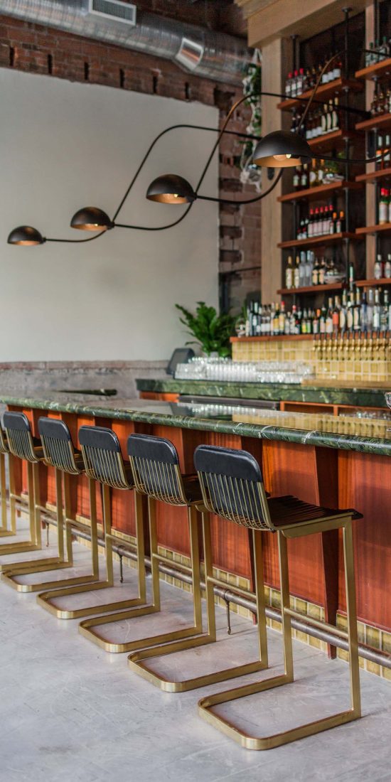Gold and black bar chairs facing a green bar with a tall liquor wall in background