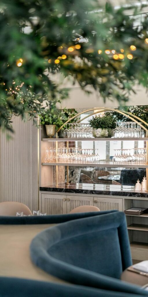 Blue velvet seating booth in the foreground and shelves with upside down wine glasses and plants in the background