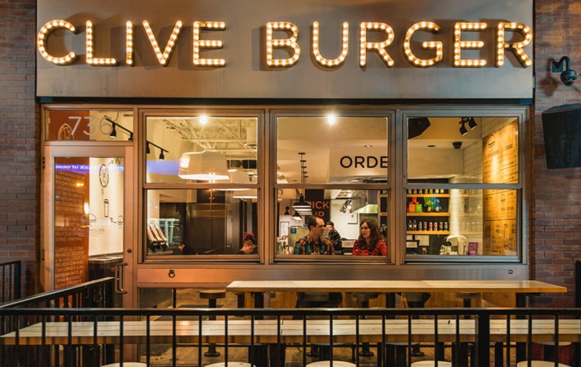 Outdoor seating patio with a large window and people inside, a lit up sign that says Clive Burger above