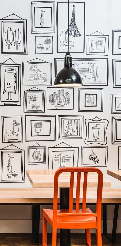An orange chair facing a wooden table and bench with sketches on the wall in the background