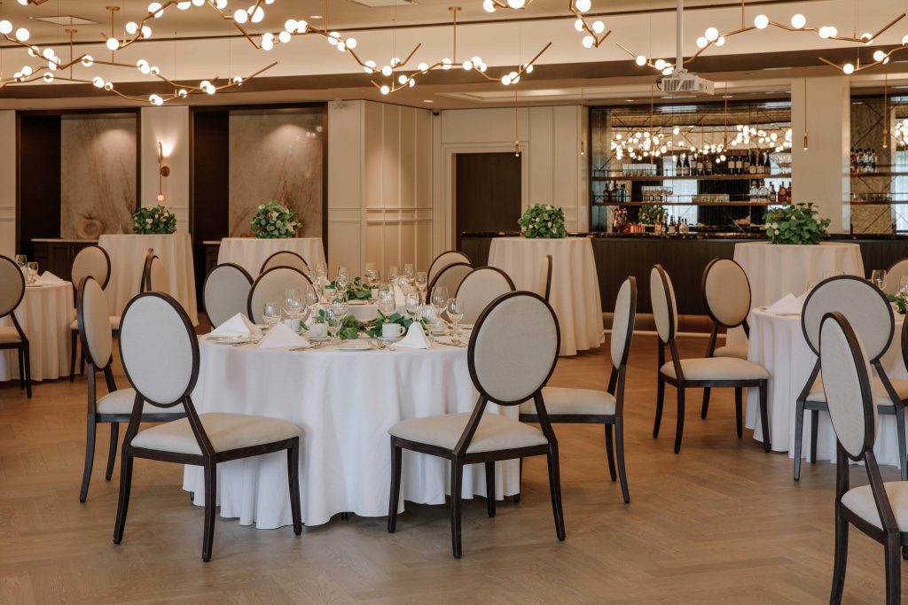 Tables draped with table cloths are set for dining and have bouquets on top. Abstract light fixtures hang above