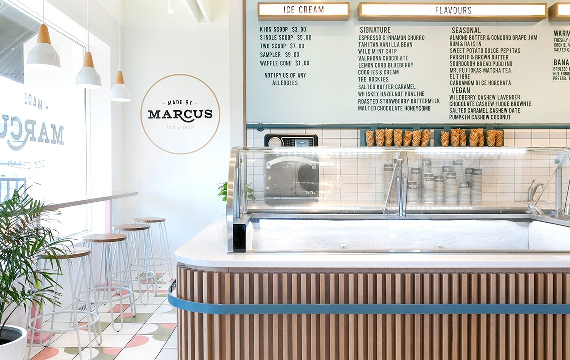 A wooden icecream counter below an icecream flavours sign next to a wall painting that says Made by Marcus
