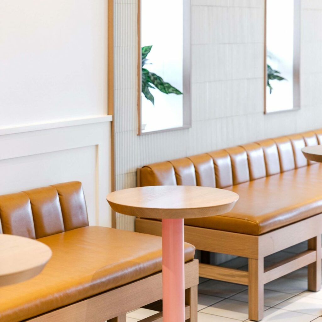 A table top on a pink stand in front of a leather seating bench with windows in the background