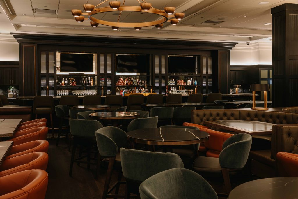 Round black marble tables are surrounded by green velvet chairs with leather seating booths on either side. In the background is a bar with tvs above liquor shelves