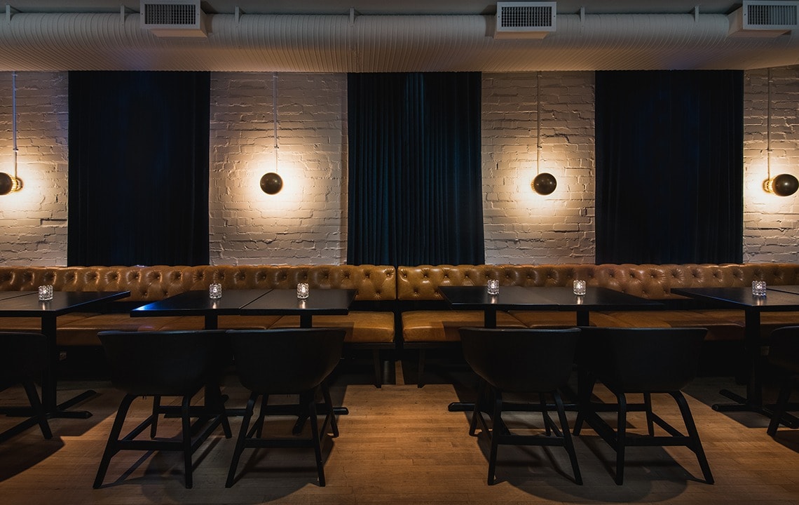 Black chairs are facing black tables with a candle on top and tan leather seating benches. A white brick wall and navy drapes are in the background