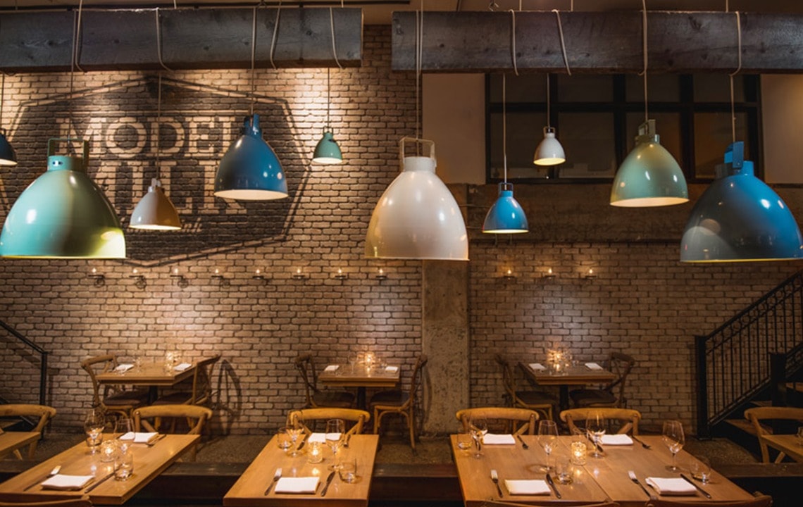 Different colour and size lamps are hanging from a wooden beam above tables set for dining