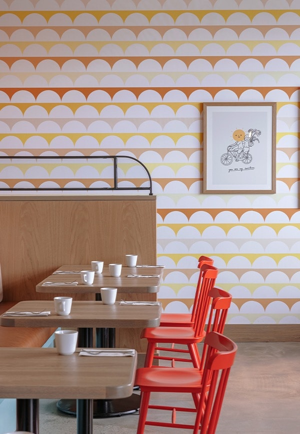 Orange chairs facing set dining tables are in front of a colourful and patterened wall with a portrait on the wall