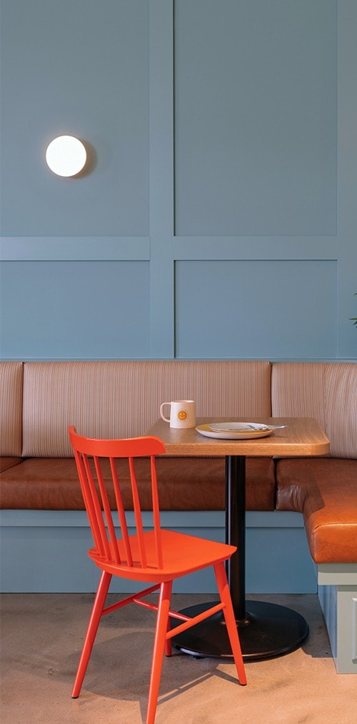 An orange chair sits in front of a table with a coffee cup and plate on top. A seating bench against a blue wall is in the background