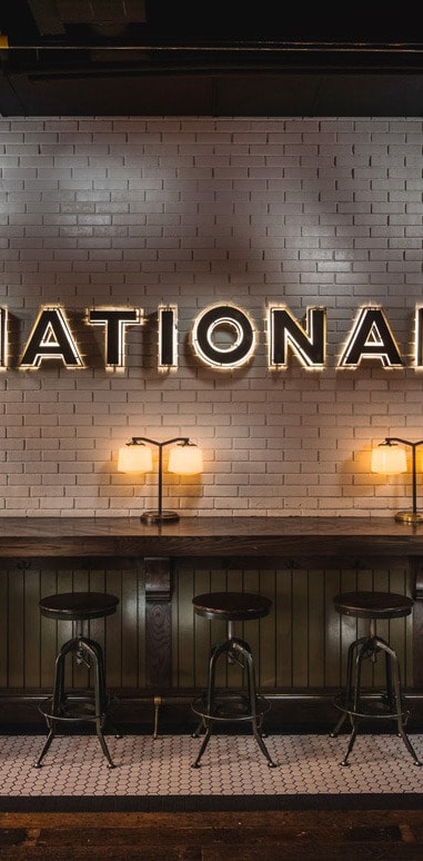 A lit up sign that says National is on a white brick wall hanging above a long wooden bar top and black bar stools