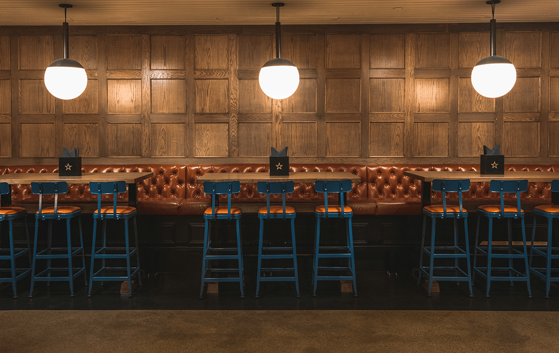 Blue and orange bar chairs face wooden bar tables and leather seating bench with globe shaped lighting fixtures hanging above the tables