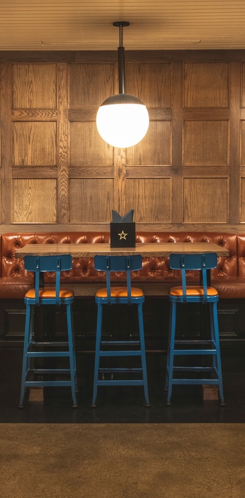 Blue and orange bar chairs face a wooden bar table with a menu on top and a circular lighting fixture hanging above