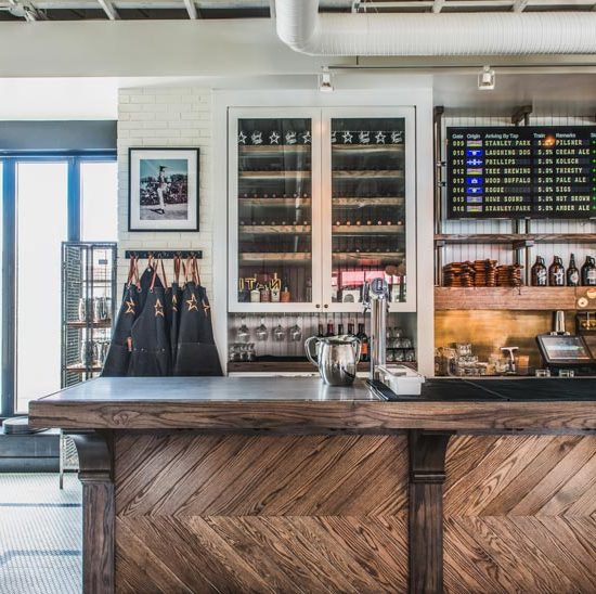 A wooden bar counter in the foreground and hanging aprons, a wine cooler, and beer menus in the background