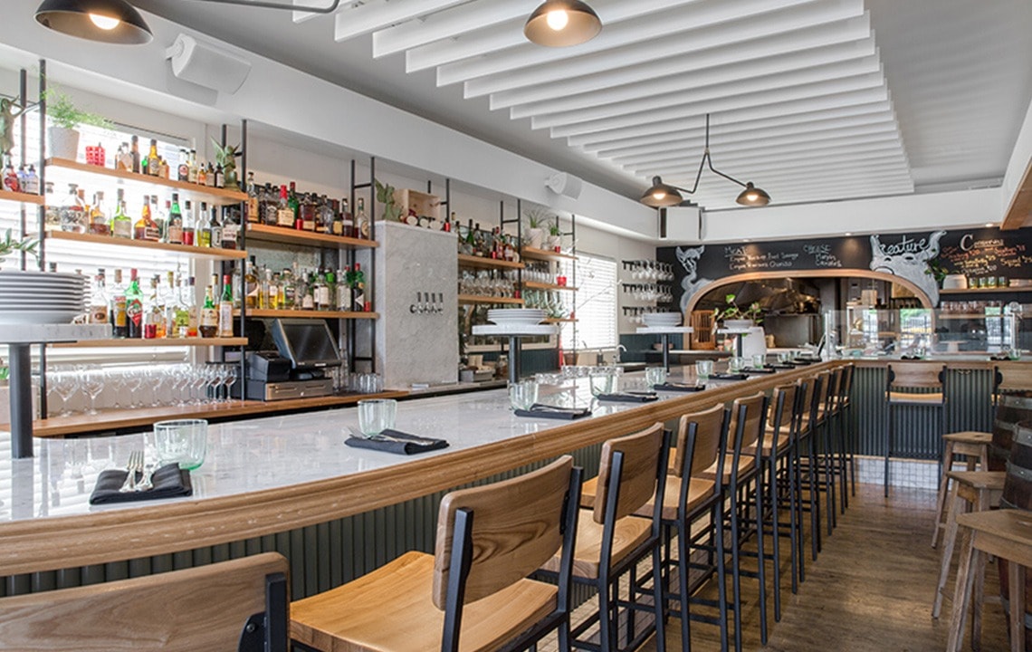 Wooden bar chairs surrounding a bar top are facing a bar with shelves that are filled with liquor bottles