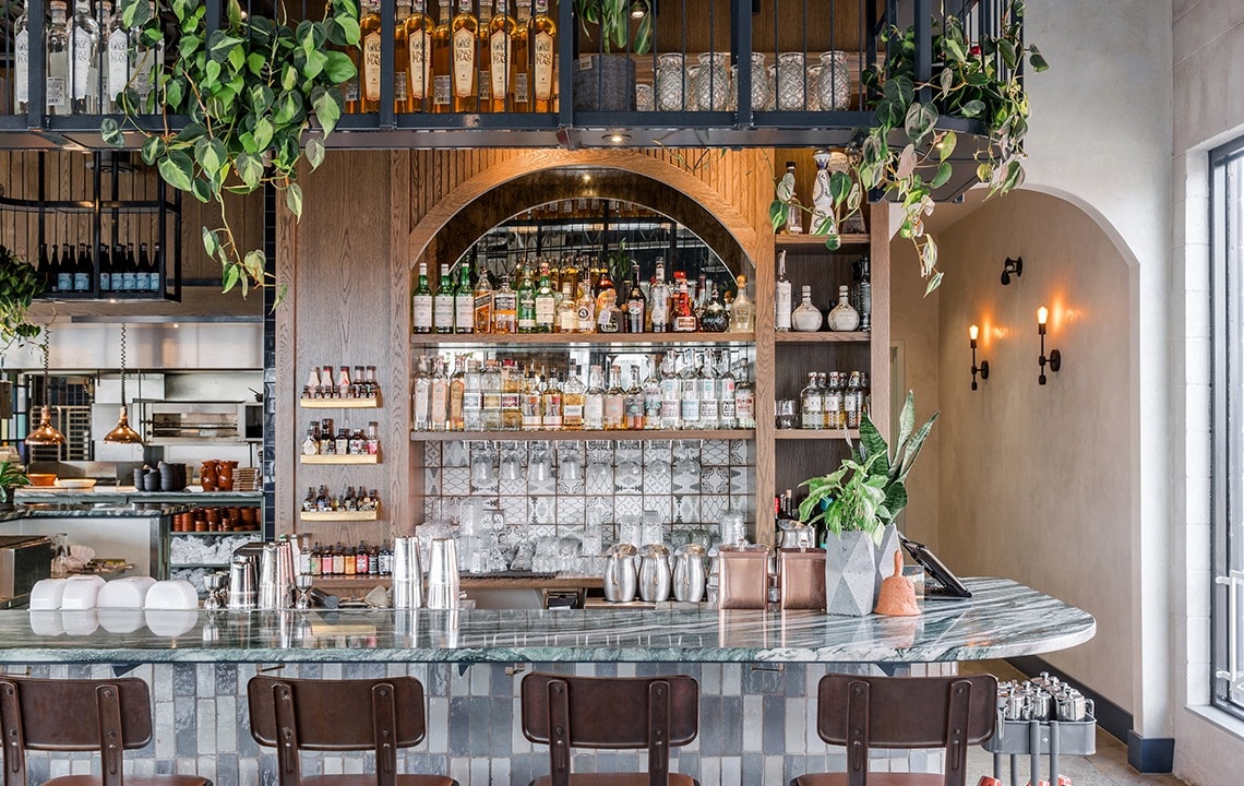 Brown bar stools in front of a marble bar top with shelves, liquor bottles and hanging plants in the background