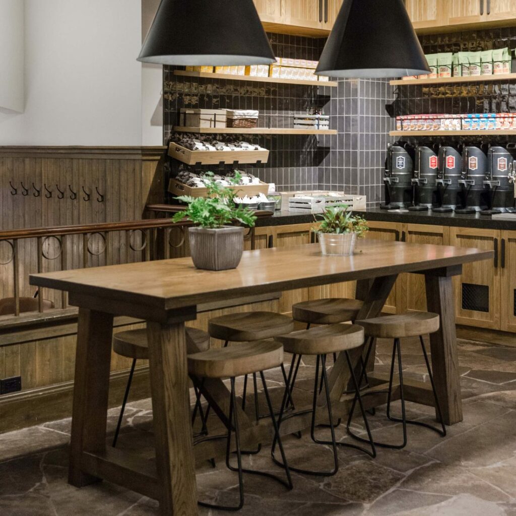 Wooden stools underneath a long wooden table with plants on top underneath back hanging lights