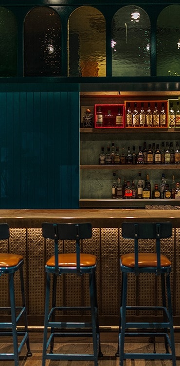 Blue bar stools sit underneath a wooden bar top with a bar with shelves filled with liquor bottles in the back