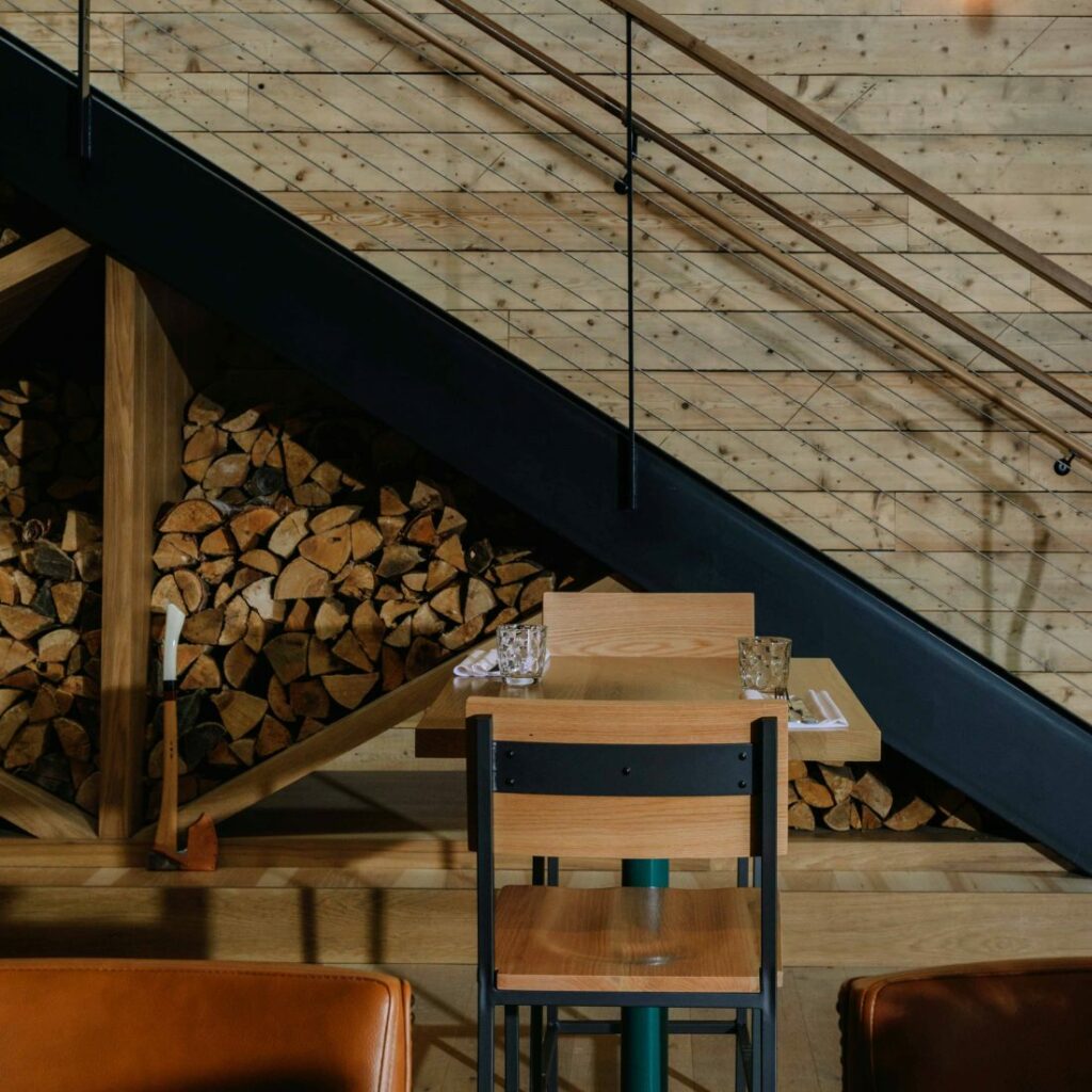 Black and wood chairs are arranged around a set table with stacks of wood tucked underneath stairs are in the background