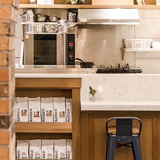 A black bar stool sits underneath a white counter top next to a shelf holding coffee bags in the foreground while industrial kitchen hardware are in the background