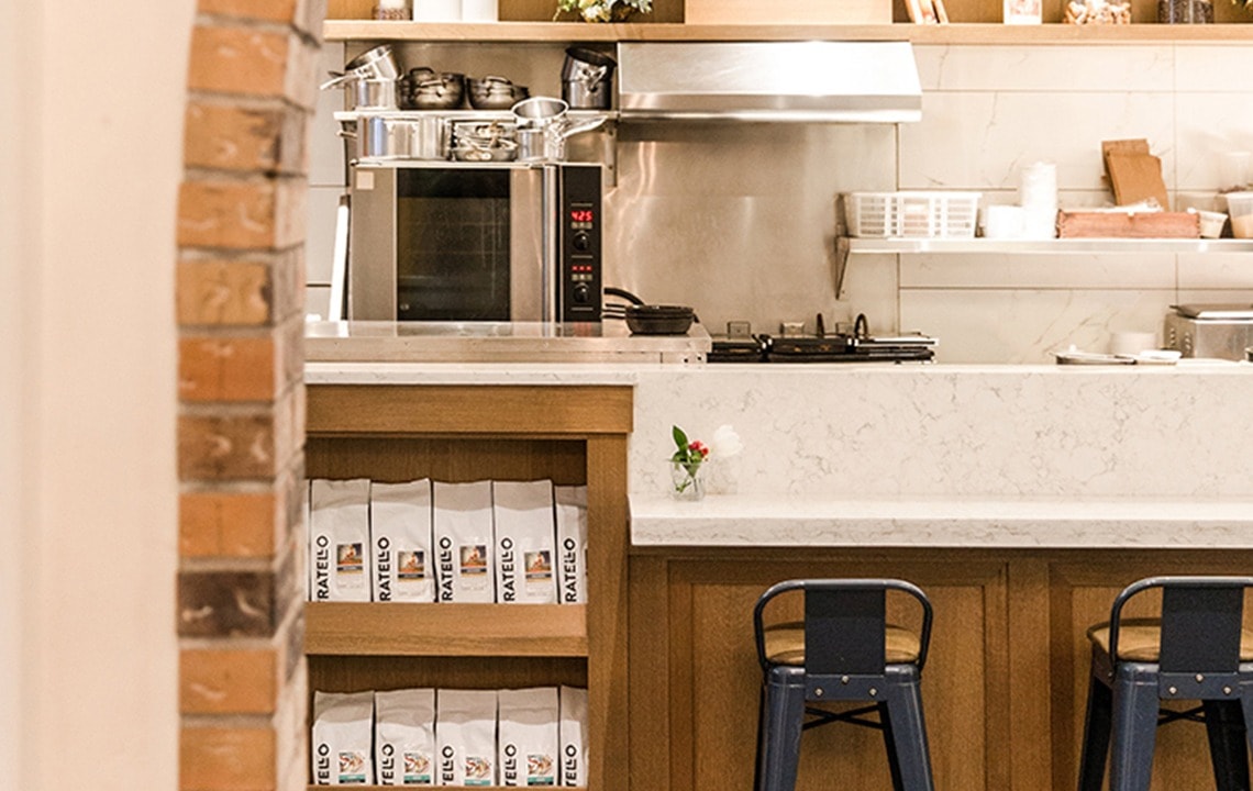 A black bar stool sits underneath a white counter top next to a shelf holding coffee bags in the foreground while industrial kitchen hardware are in the background