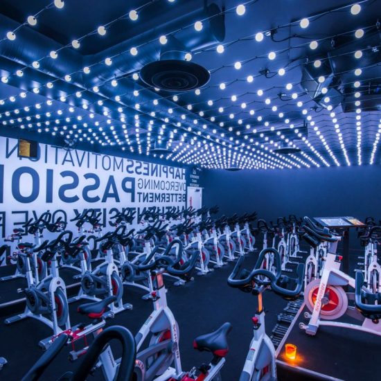 A room filled with stationary bikes are underneath liines of bright white lights. On the back wall are motivational quotes painted on top