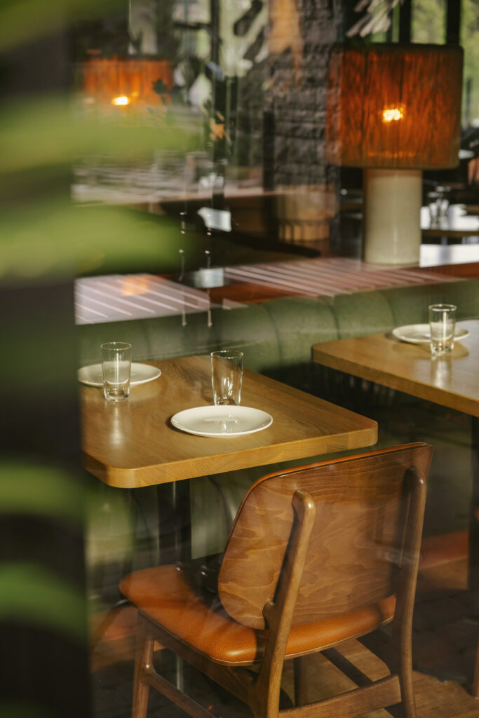 An artistic view through a window and plant leaves of a restaurant table. On one side of the table is a wooden chair with a leather seat. On the other side is a green fabric banquette.