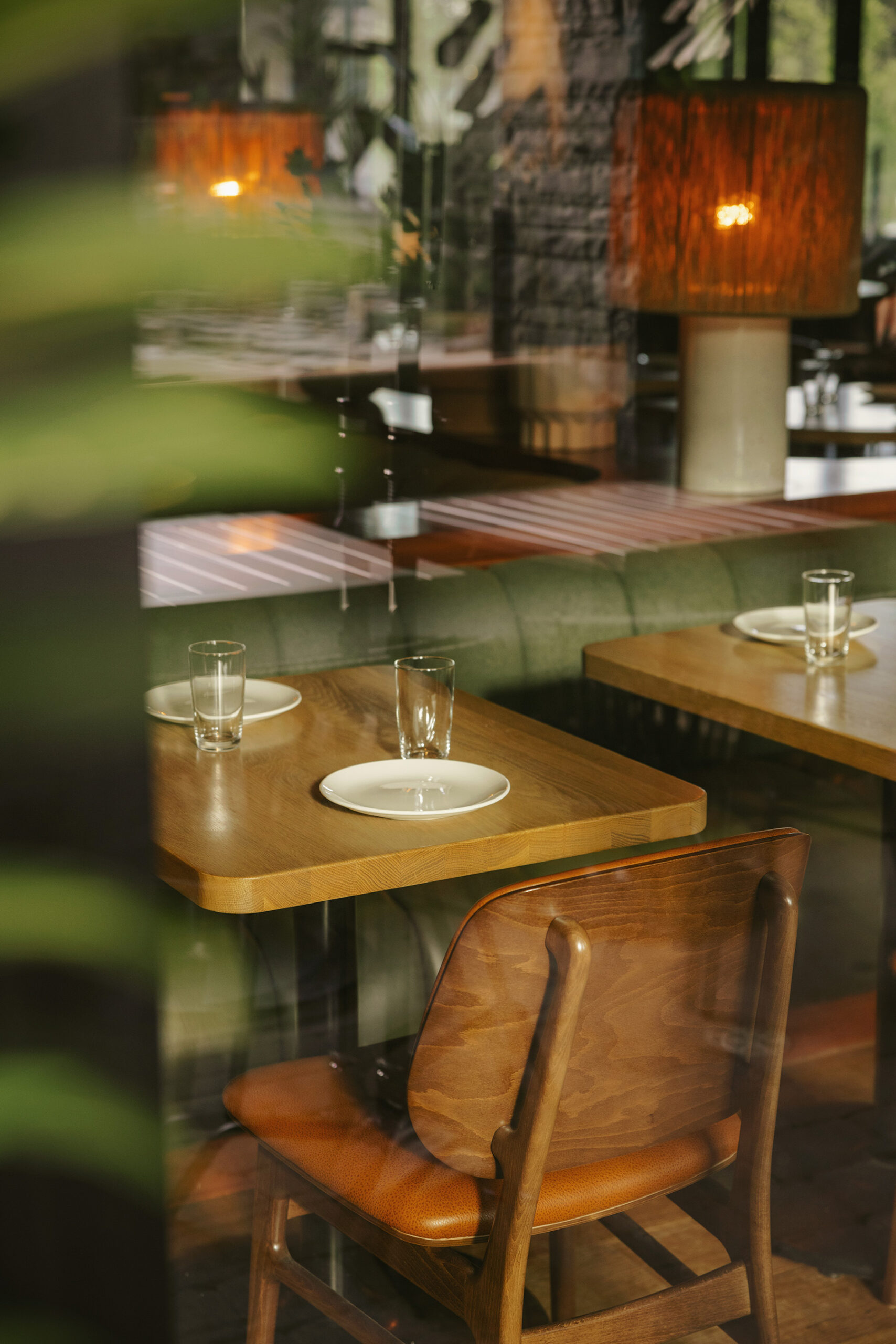 An artistic view through a window and plant leaves of a restaurant table. On one side of the table is a wooden chair with a leather seat. On the other side is a green fabric banquette.