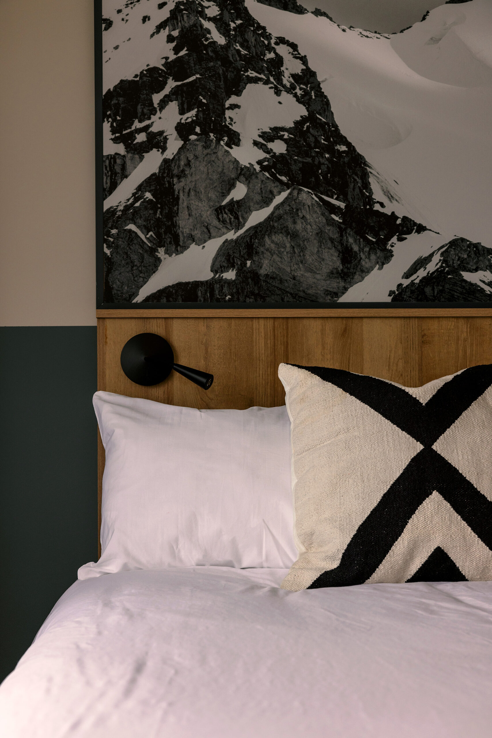 Cropped photo of a bed with white linens and a throw pillow with a black X on it. There is a large black and white mountain photograph on the wall above the wooden headboard. The walls are green.
